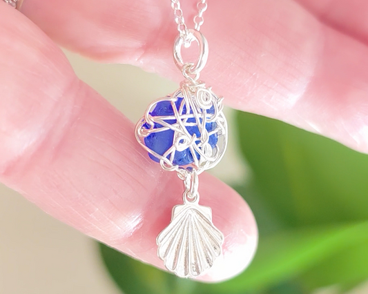 Silver Wire Wrapped Sapphire cobalt blue sea glass pendant with dangling shell on rolo chain in hand 