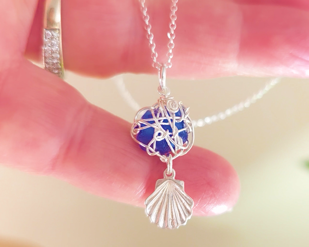 Silver Wire Wrapped Sapphire cobalt blue sea glass pendant with dangling shell on rolo chain in hand 