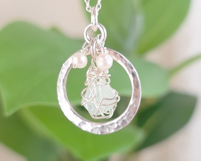 Round Circle Silver Pendant with wire wrapped Aqua Blue Beach Glass and two small white pearls on chain with blurred green background 