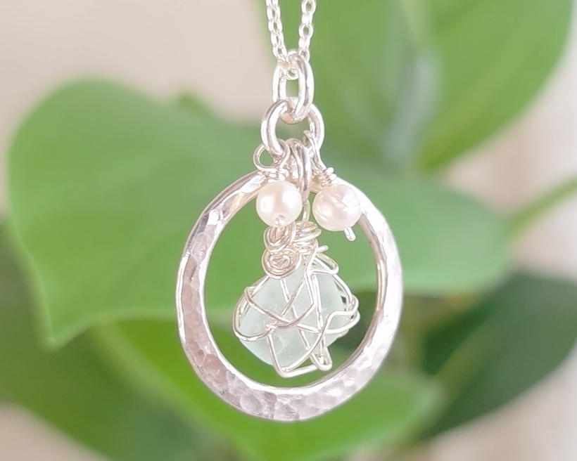 Round Circle Silver Pendant with wire wrapped Aqua Blue Beach Glass and two small white pearls on chain with blurred green background 