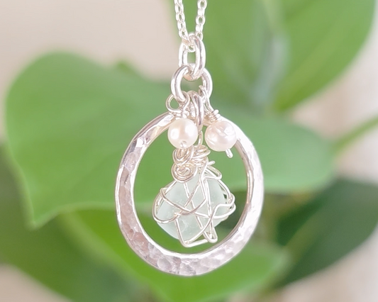 Round Circle Silver Pendant with wire wrapped Aqua Blue Beach Glass and two small white pearls on chain with blurred green background 