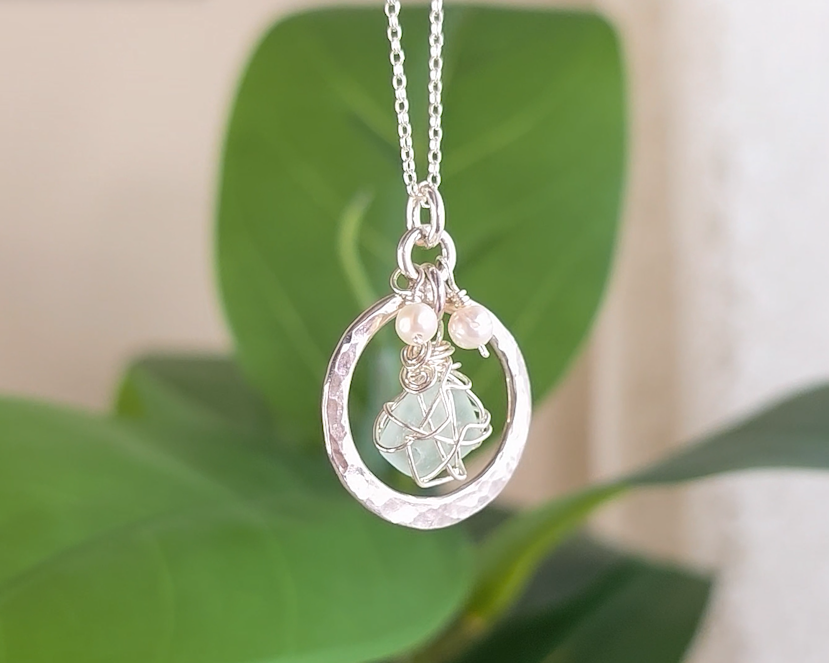 Round Circle Silver Pendant with wire wrapped Aqua Blue Beach Glass and two small white pearls on chain with blurred green background 