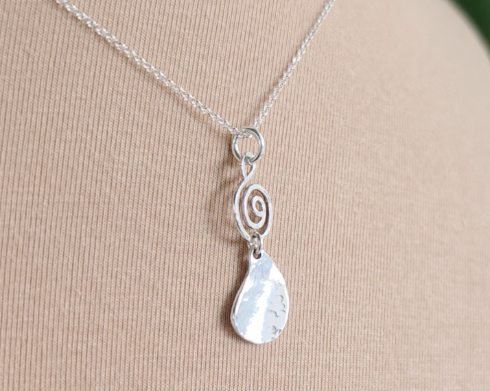 Handmade sterling silver pendant with Celtic Coil on top and drop shaped pendant dangling below on silver chain with beige background 