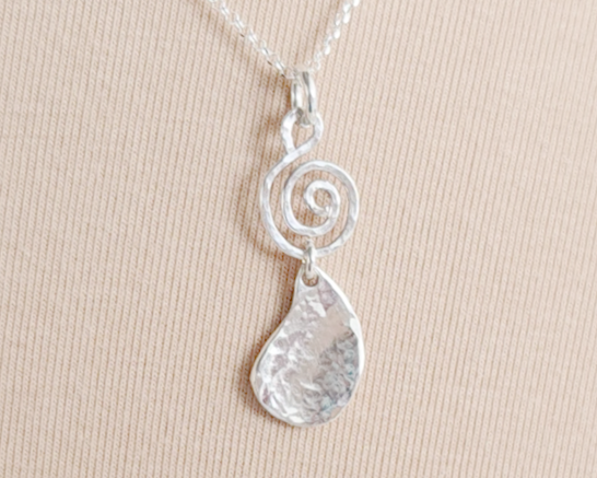 Handmade sterling silver pendant with Celtic Coil on top and drop shaped pendant dangling below on silver chain with beige background 