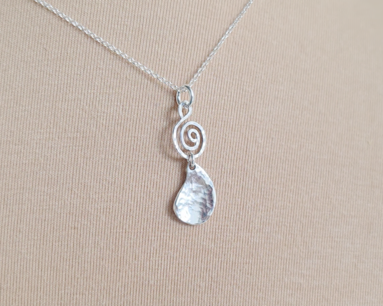 Handmade sterling silver pendant with Celtic Coil on top and drop shaped pendant dangling below on silver chain with beige background 
