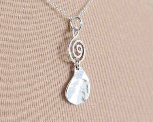 Handmade sterling silver pendant with Celtic Coil on top and drop shaped pendant dangling below on silver chain with beige background 