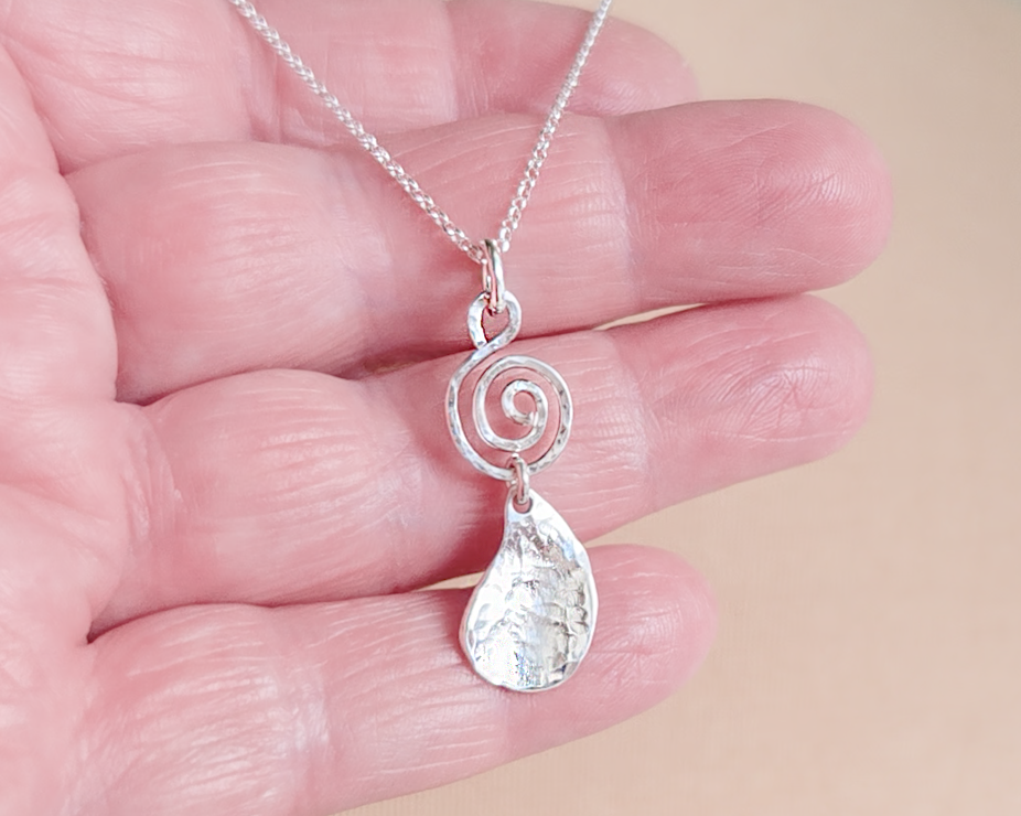 Handmade sterling silver pendant with Celtic Coil on top and drop shaped pendant dangling below on silver chain in hand