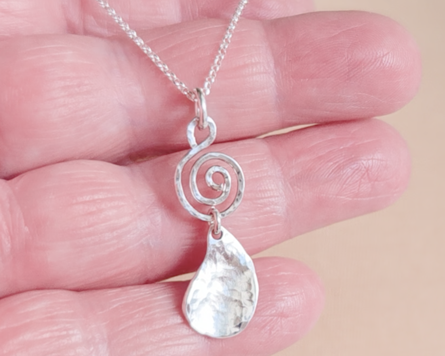 Handmade sterling silver pendant with Celtic Coil on top and drop shaped pendant dangling below on silver chain in hand