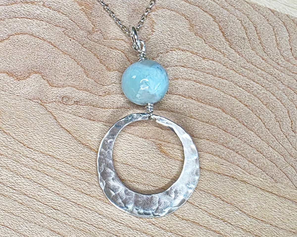 Handmade round sterling silver pendant with wire wrapped round pale blue larimar stone dangling on chain displayed on wood background 