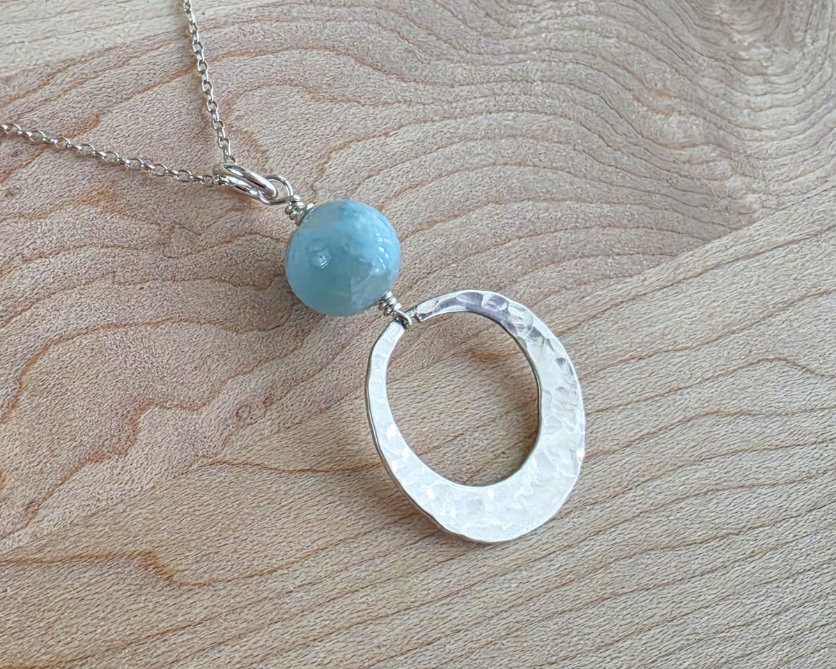Handmade round sterling silver pendant with wire wrapped round pale blue larimar stone dangling on chain displayed on wood background 