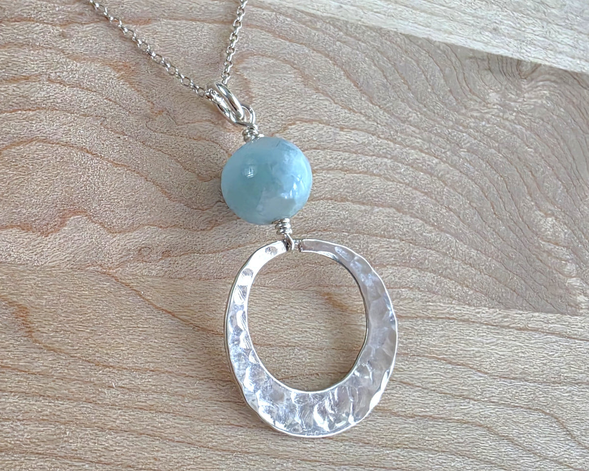 Handmade round sterling silver pendant with wire wrapped round pale blue larimar stone dangling on chain displayed on wood background 