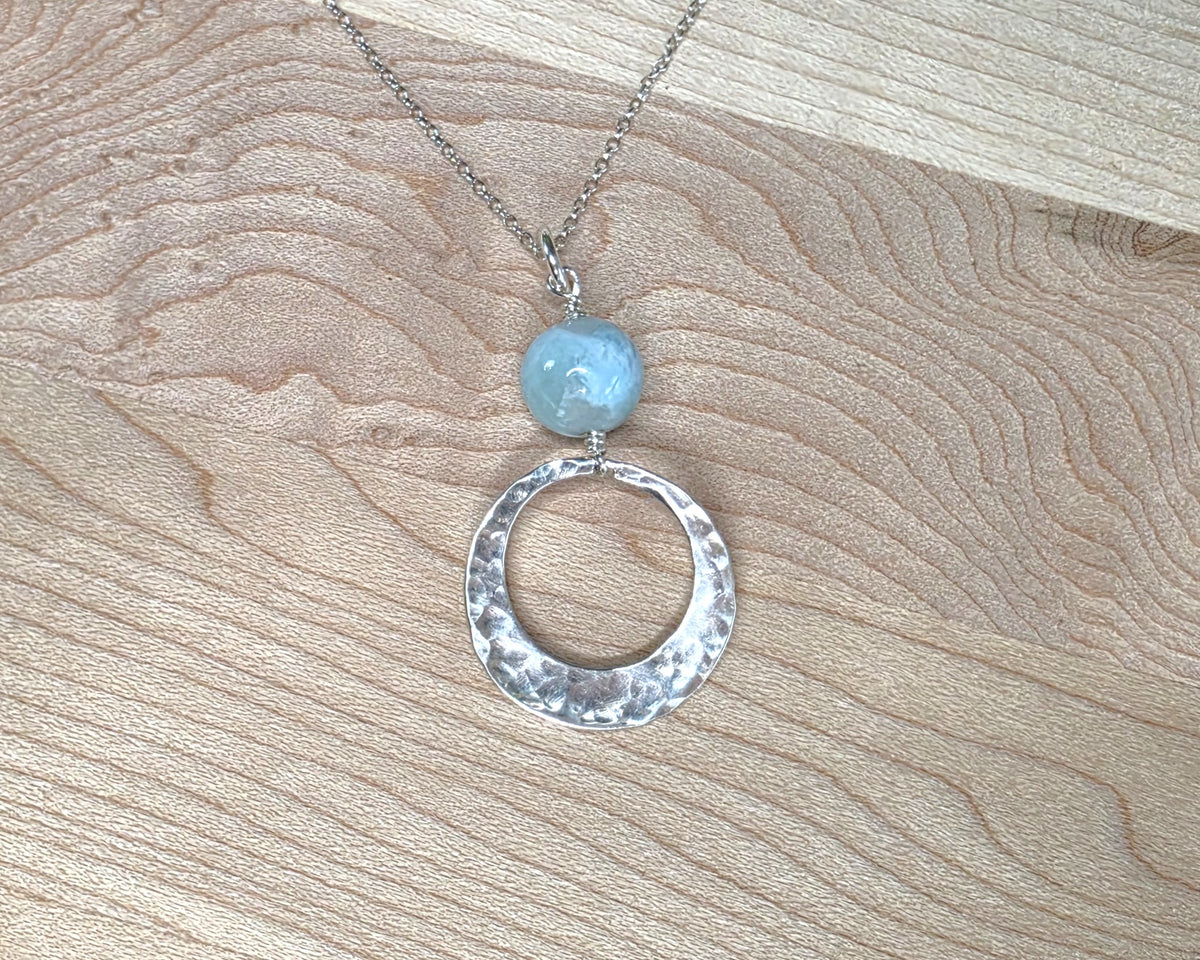 Handmade round sterling silver pendant with wire wrapped round pale blue larimar stone dangling on chain displayed on wood background 