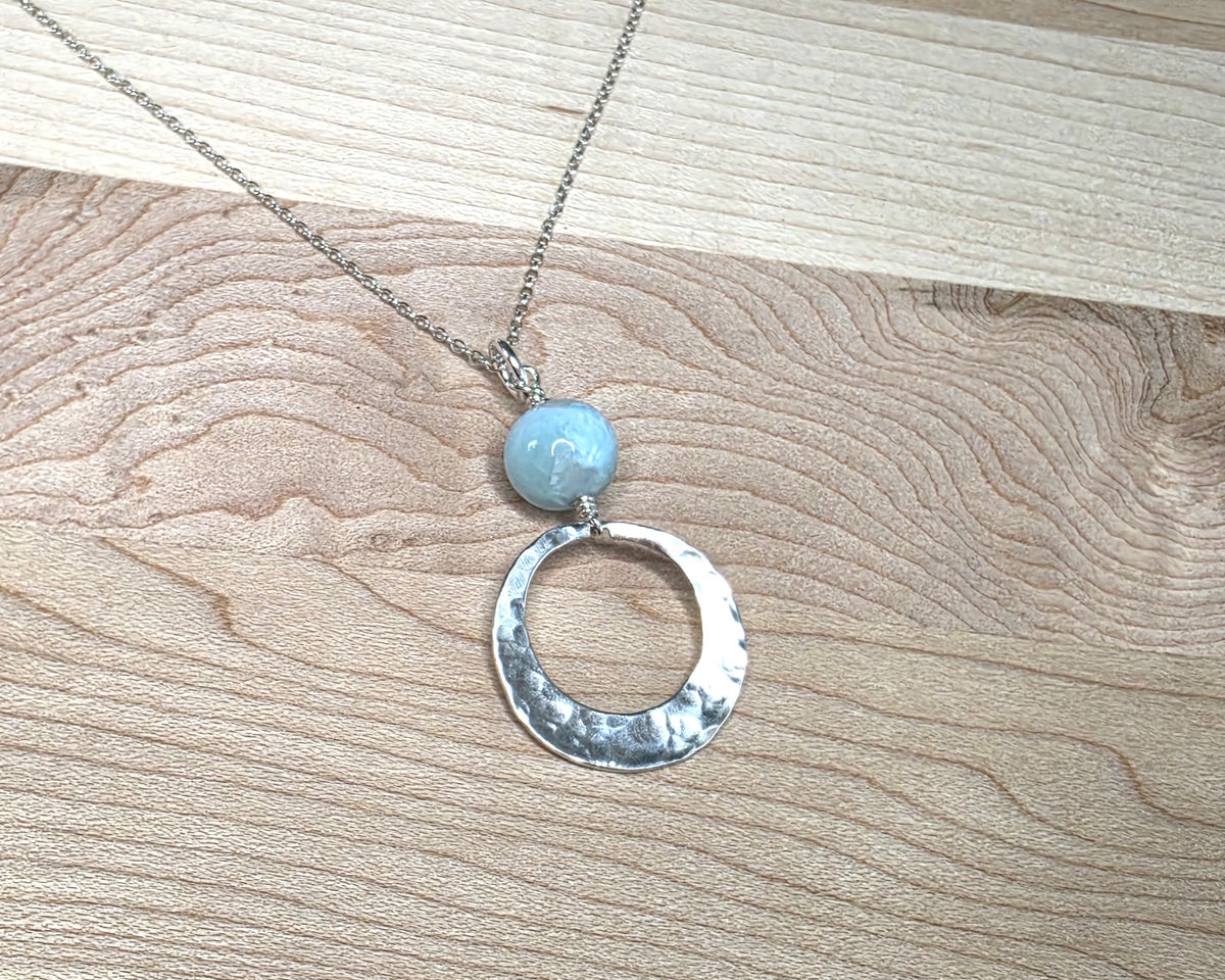 Handmade round sterling silver pendant with wire wrapped round pale blue larimar stone dangling on chain displayed on wood background 