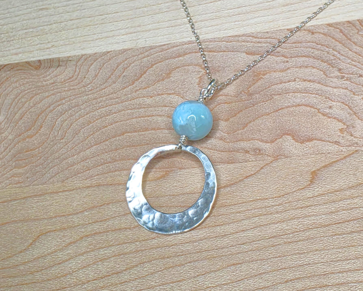 Handmade round sterling silver pendant with wire wrapped round pale blue larimar stone dangling on chain displayed on wood background 