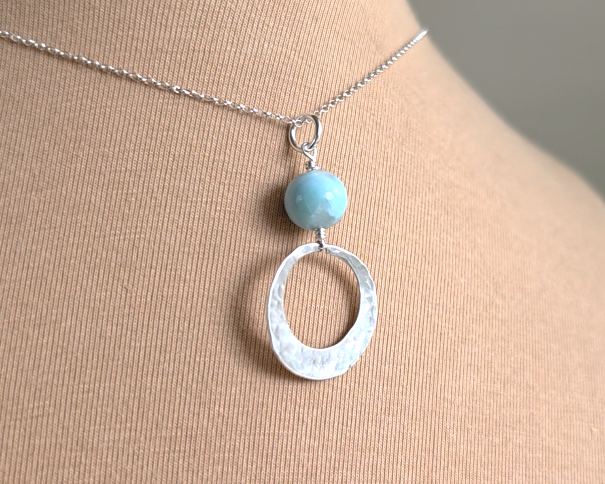 Handmade round sterling silver pendant with wire wrapped round pale blue larimar stone dangling on chain displayed on beige display bust