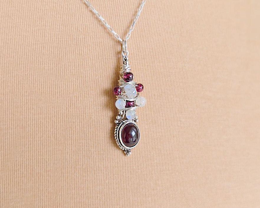 Handmade Boho style wire wrapped pendant with oval shape deep red Garnet gemstone wrapped with sterling silver wire small round garnets and faceted Moonstones on chain with beige cloth background 