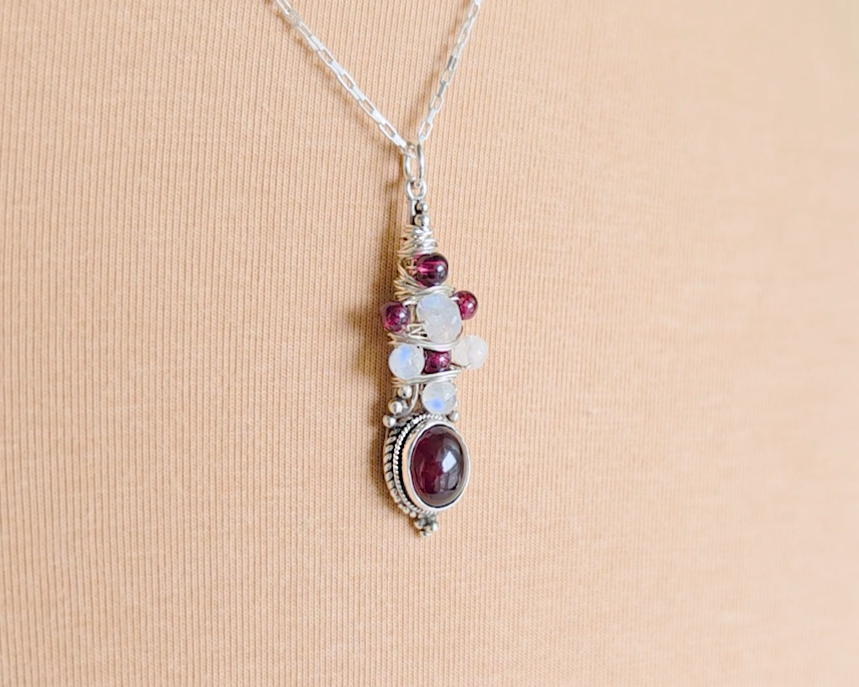 Handmade Boho style wire wrapped pendant with oval shape deep red Garnet gemstone wrapped with sterling silver wire small round garnets and faceted Moonstones on chain with beige cloth background 