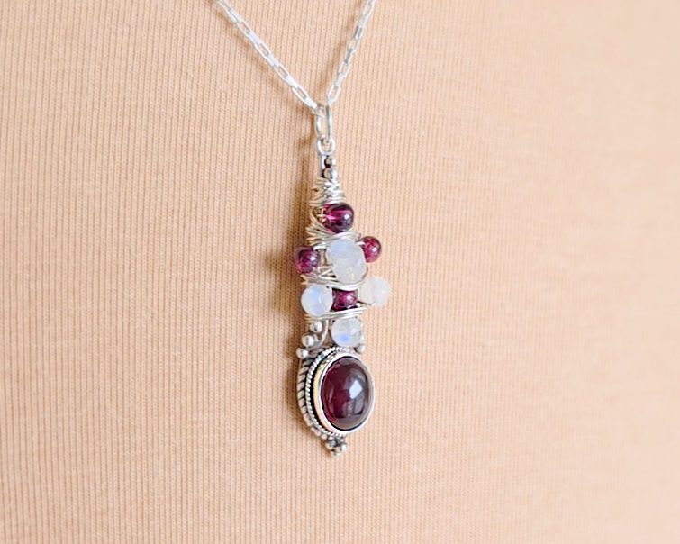 Handmade Boho style wire wrapped pendant with oval shape deep red Garnet gemstone wrapped with sterling silver wire small round garnets and faceted Moonstones on chain with beige cloth background 