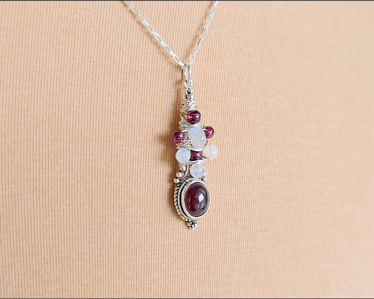 Handmade Boho style wire wrapped pendant with oval shape deep red Garnet gemstone wrapped with sterling silver wire small round garnets and faceted Moonstones on chain with beige cloth background 