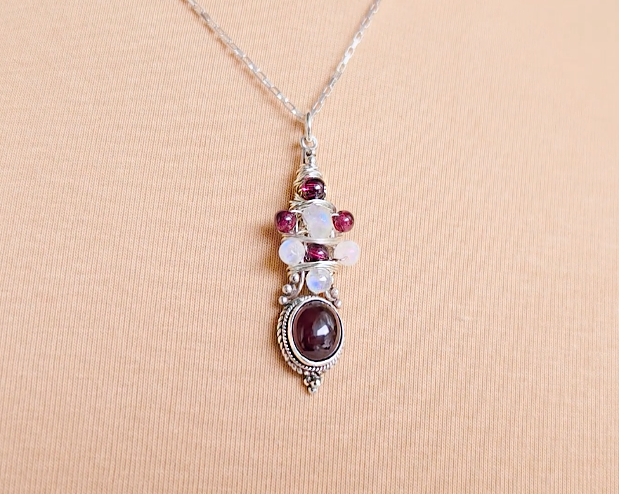 Handmade Boho style wire wrapped pendant with oval shape deep red Garnet gemstone wrapped with sterling silver wire small round garnets and faceted Moonstones on chain with beige cloth background 
