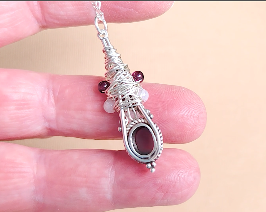 Back of Handmade Boho style wire wrapped pendant with oval shape deep red Garnet gemstone wrapped with sterling silver wire small round garnets and faceted Moonstones in hand 