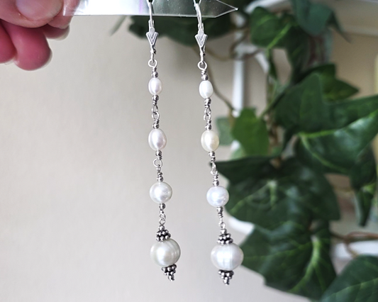 Long art deco style pearl earrings, white Freshwater Cultured Pearls, accented with fancy Sterling Silver beads on lever back earrings hooks on white background with leaves