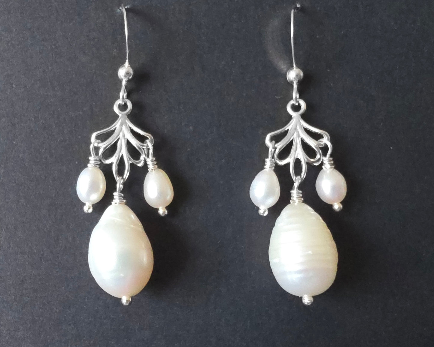 Sterling Silver Large white drop shaped pearl earrings with two small drop shaped pearls dangling on either side from and open leaf like design, the earrings dangle from french earrings hooks, displayed on black background. 