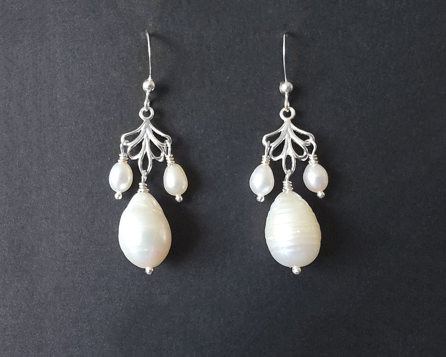 Sterling Silver Large white drop shaped pearl earrings with two small drop shaped pearls dangling on either side from and open leaf like design, the earrings dangle from french earrings hooks, displayed on black background. 