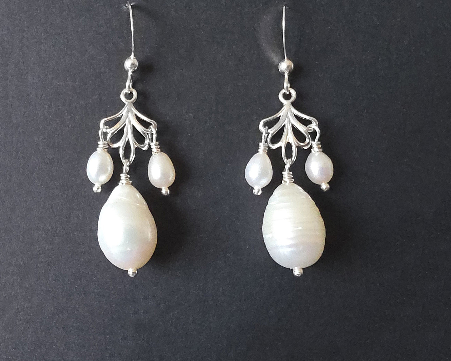 Sterling Silver Large white drop shaped pearl earrings with two small drop shaped pearls dangling on either side from and open leaf like design, the earrings dangle from french earrings hooks, displayed on black background. 