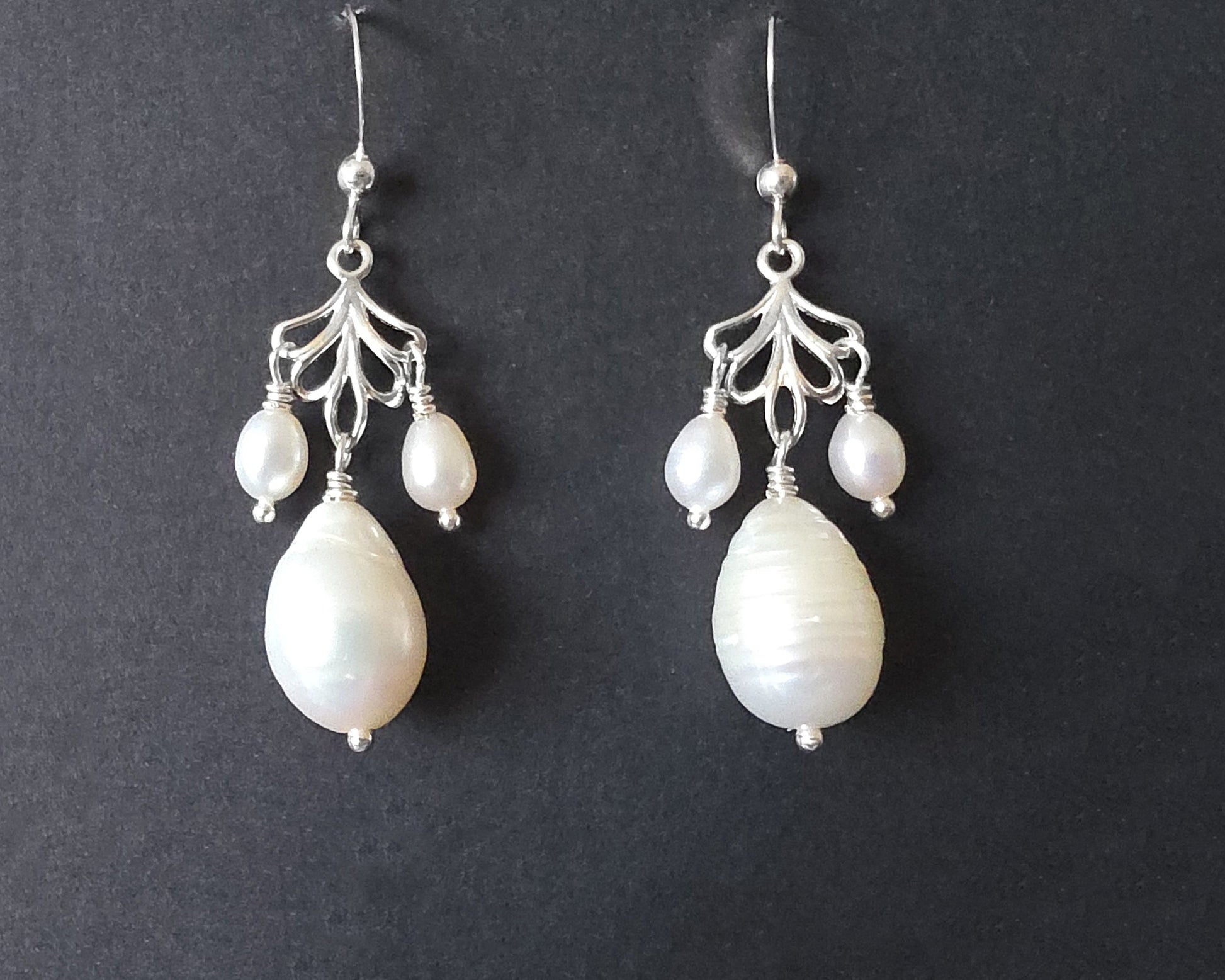 Sterling Silver Large white drop shaped pearl earrings with two small drop shaped pearls dangling on either side from and open leaf like design, the earrings dangle from french earrings hooks, displayed on black background. 