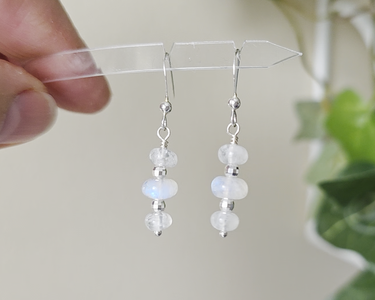 Sterling Silver Dangle Earrings with Three milky white Moonstones with blue and Sterling Silver sparkly beads dangling from silver French earring hooks held Infront of green leaf background 
