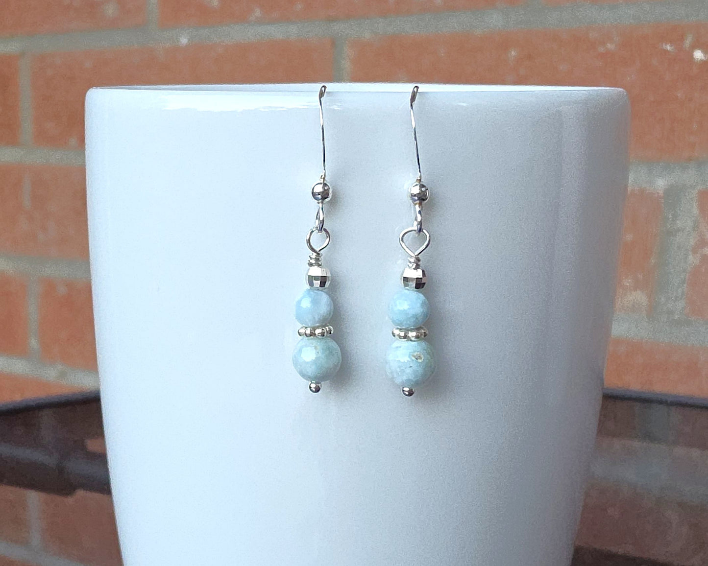 A pair of pale blue gemstone sterling silver dangle earrings on french earring hooks on white cup with brick wall in background 