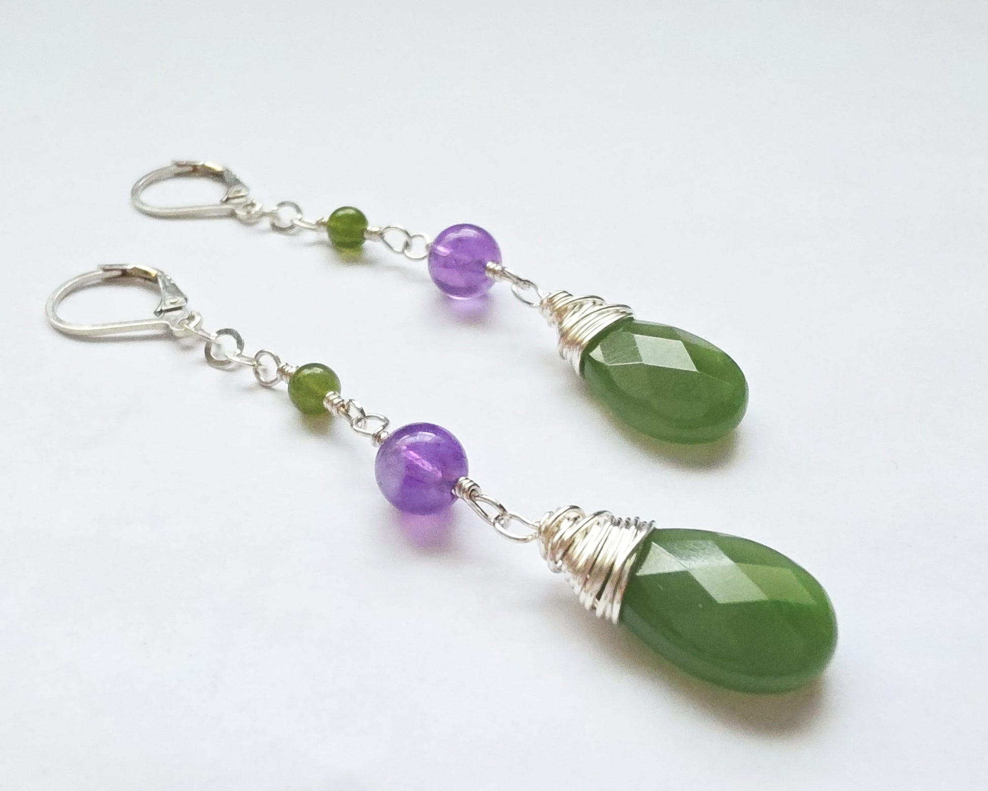 A pair of handmade long dangle drop earrings with green and purple stones, a drop shape Green Jade, wire wrapped with Amethyst and smaller Jade beads on Lever Back earring hooks on white background