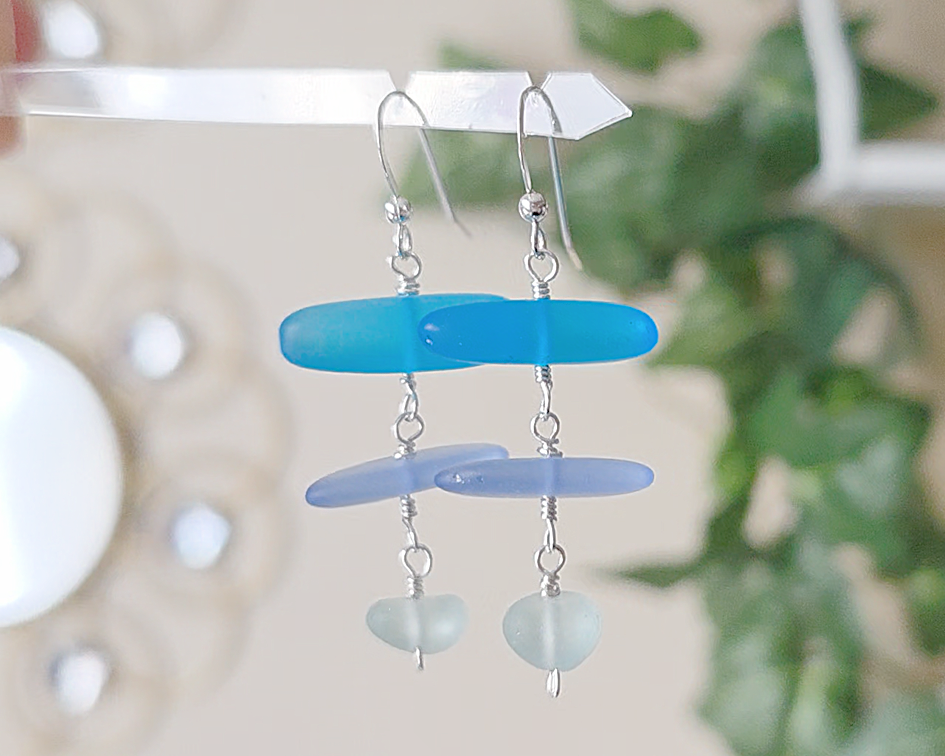 : Long Silver Sea glass earrings with three different colors of blue glass from aqua to sky blue on french style earring hooks with plants in the background 