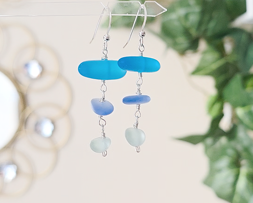 : Long Silver Sea glass earrings with three different colors of blue glass from aqua to sky blue on french style earring hooks with plants in the background 
