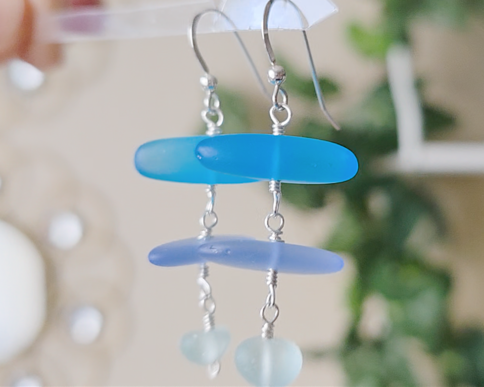 : Long Silver Sea glass earrings with three different colors of blue glass from aqua to sky blue on french style earring hooks with plants in the background 
