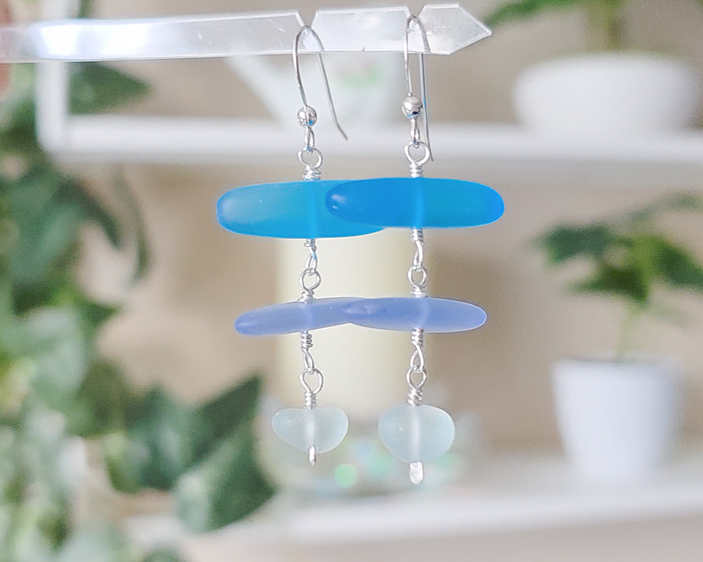 : Long Silver Sea glass earrings with three different colors of blue glass from aqua to sky blue on french style earring hooks with plants in the background 