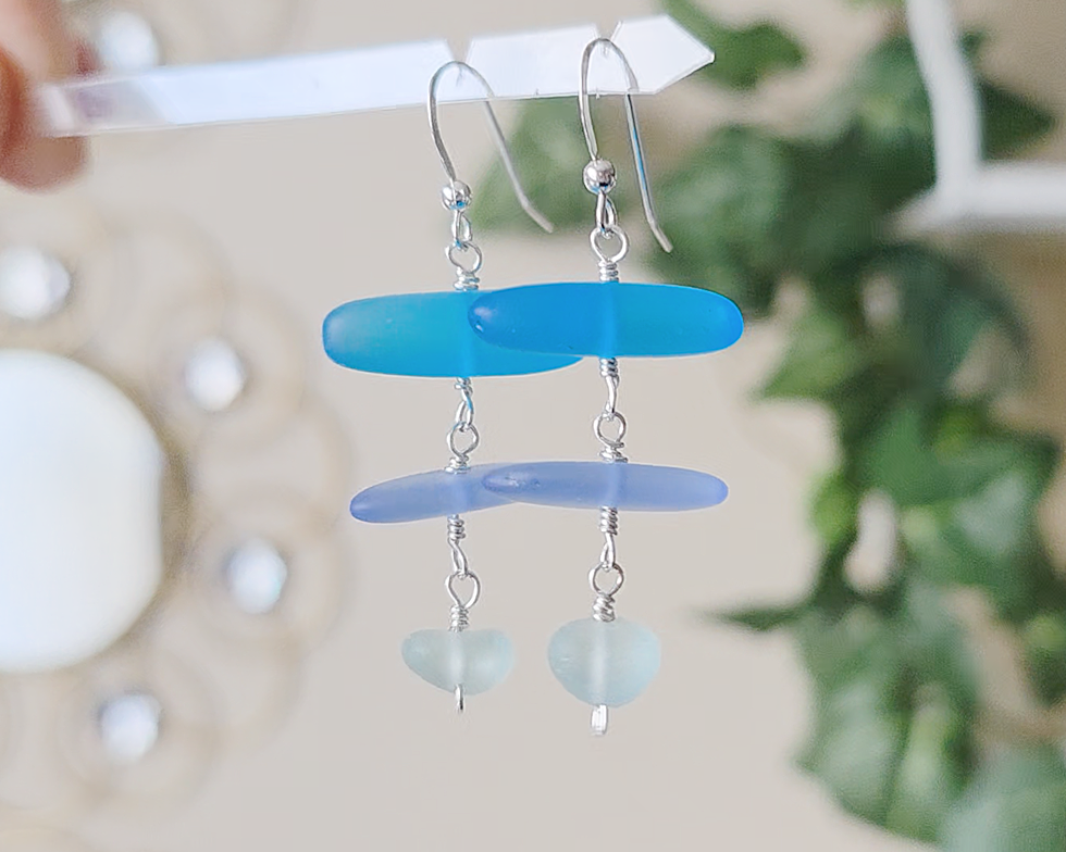 : Long Silver Sea glass earrings with three different colors of blue glass from aqua to sky blue on french style earring hooks with plants in the background 