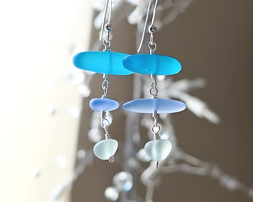 : Long Silver Sea glass earrings with three different colors of blue glass from aqua to sky blue on french style earring hooks with plants in the background 