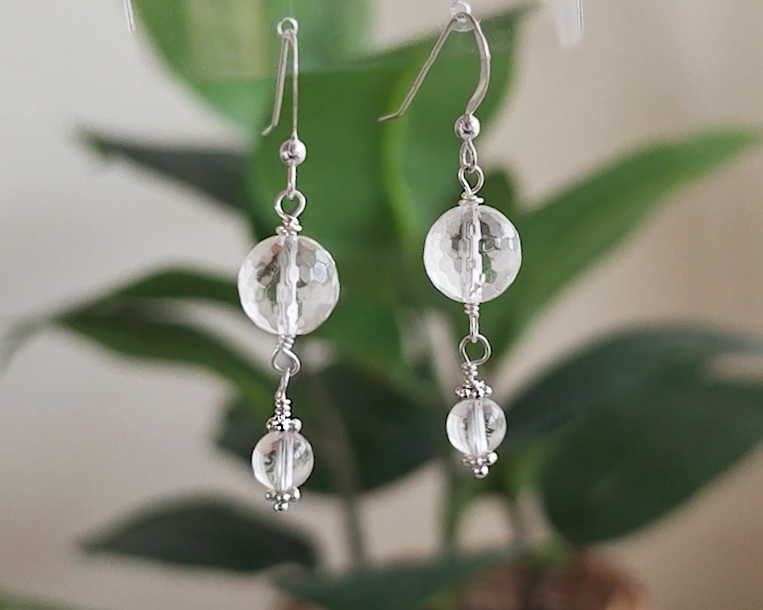 A pair of clear Quartz Crystal Earrings with faceted and polished stones with Sterling Silver beads, wire wrapped and french hooks hanging with blurry plants in background 