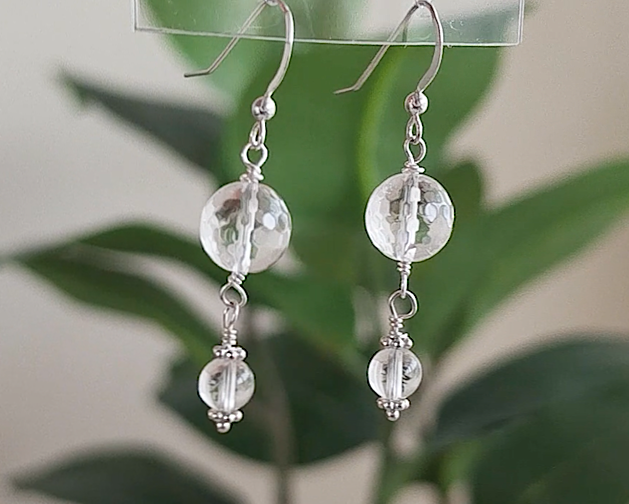 A pair of clear Quartz Crystal Earrings with faceted and polished stones with Sterling Silver beads, wire wrapped and french hooks hanging with blurry plants in background 