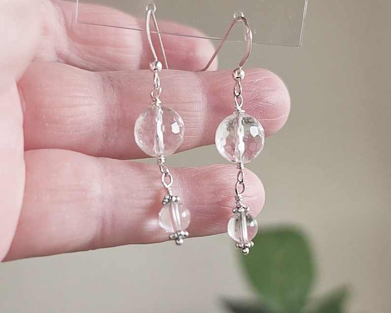 A pair of clear Quartz Crystal Earrings with faceted and polished stones with Sterling Silver beads, wire wrapped and french hooks in hand with blurry plants in background 