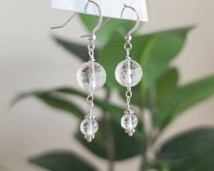 A pair of clear Quartz Crystal Earrings with faceted and polished stones with Sterling Silver beads, wire wrapped and french hooks hanging with blurry plants in background