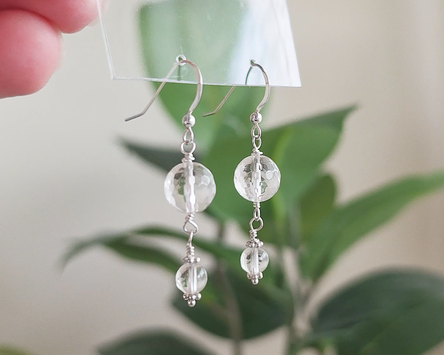 A pair of clear Quartz Crystal Earrings with faceted and polished stones with Sterling Silver beads, wire wrapped and french hooks hanging with blurry plants in background