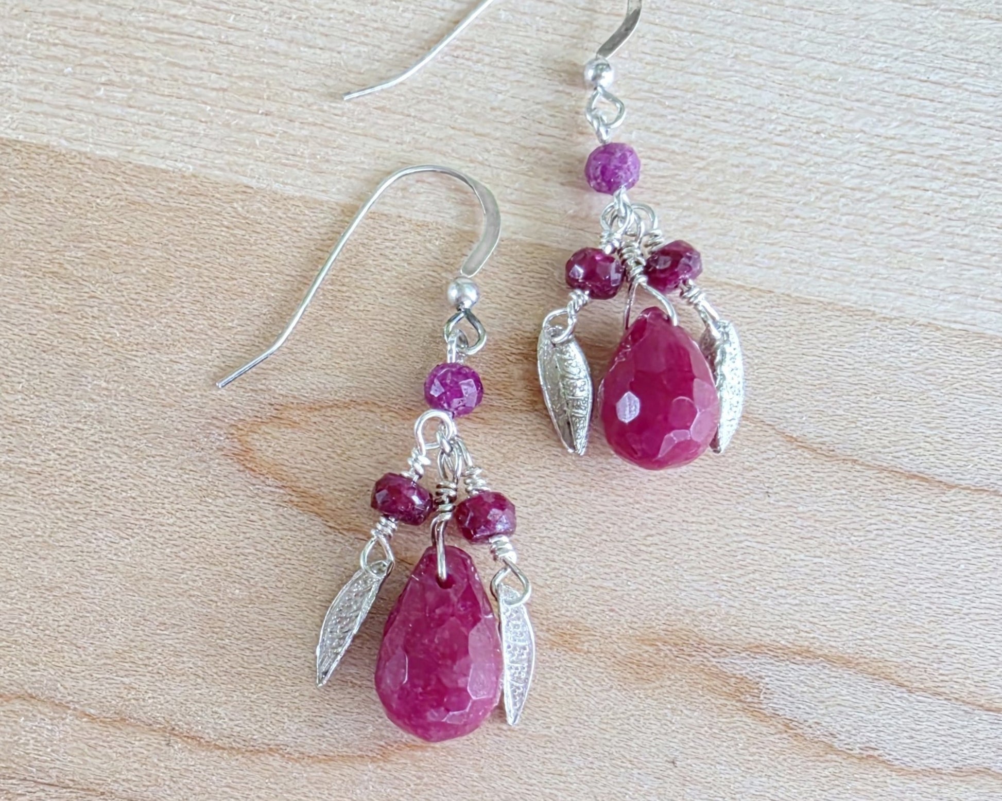A pair of handmade Ruby earrings with Sterling Silver leaf’s, large drop shape red gemstone and red rubies with french earring hooks on light color wood