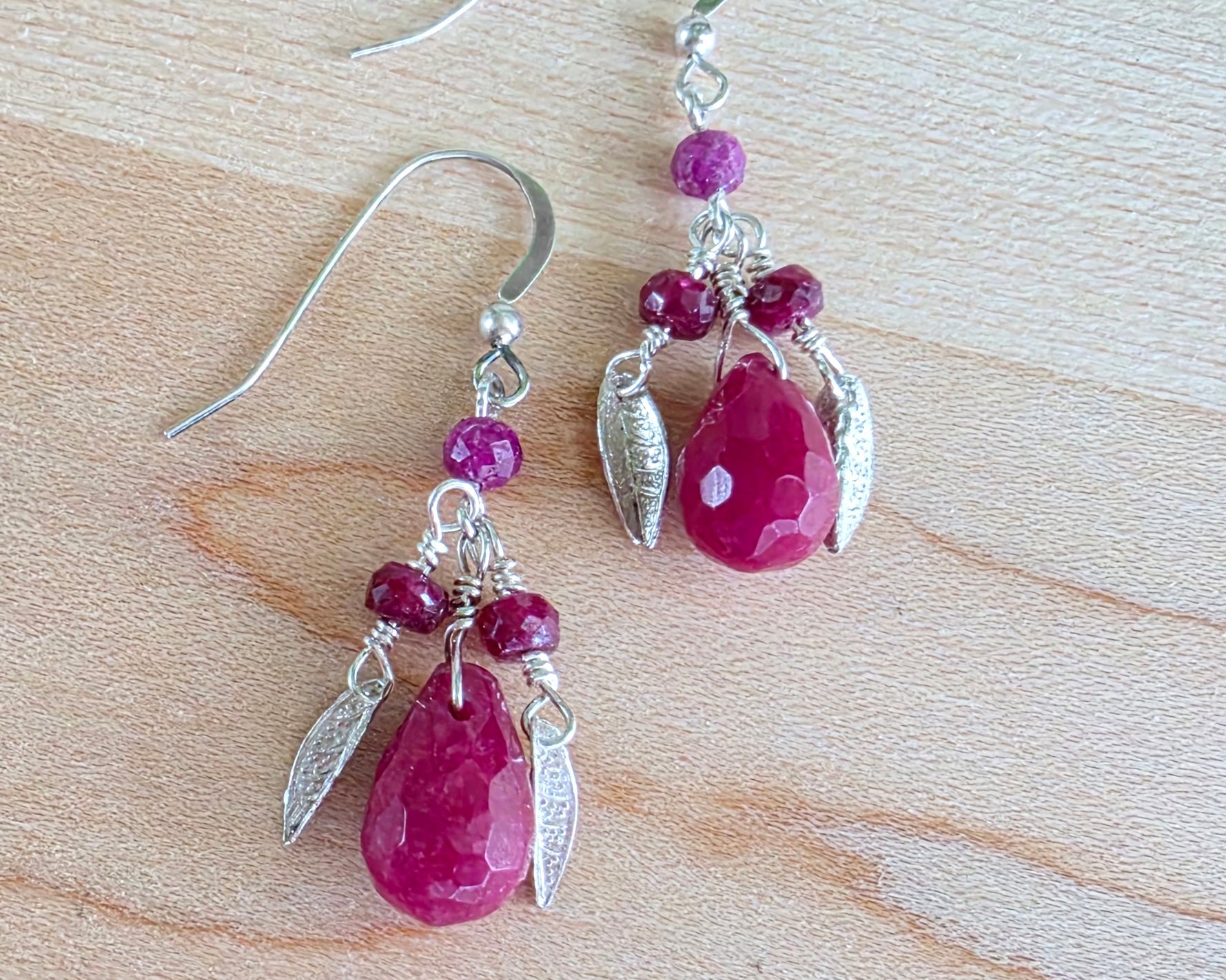 A pair of handmade Ruby earrings with Sterling Silver leaf’s, large drop shape red gemstone and red rubies with french earring hooks on light color wood