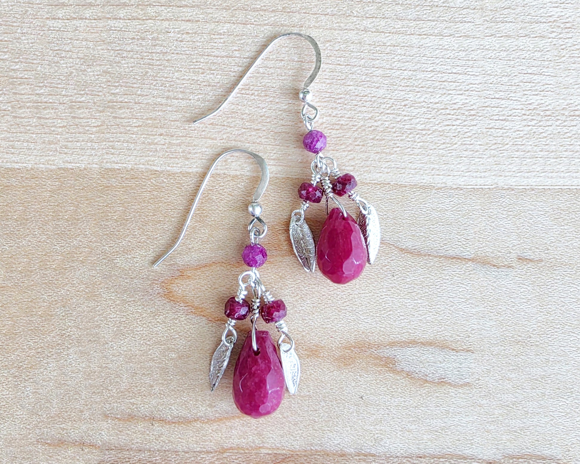 A pair of handmade Ruby earrings with Sterling Silver leaf’s, large drop shape red gemstone and red rubies with french earring hooks on light color wood