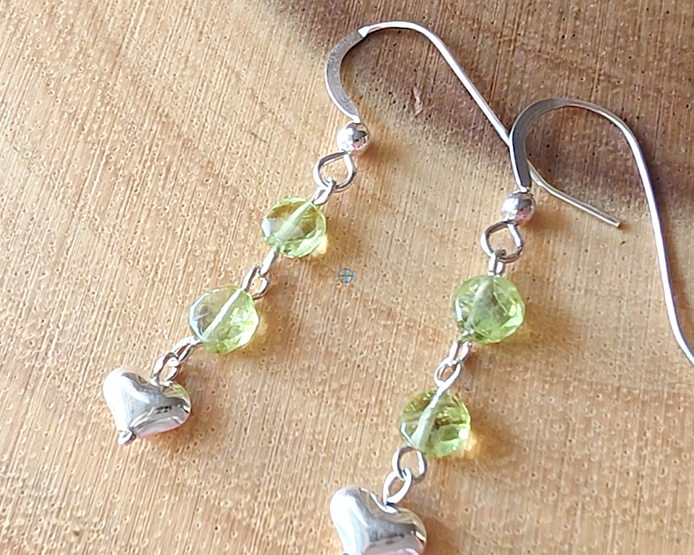 A pair of long sparkly Peridot Sterling silver earrings with two round faceted green gemstones and silver hearts on the base dangling from french earring hooks on wood 