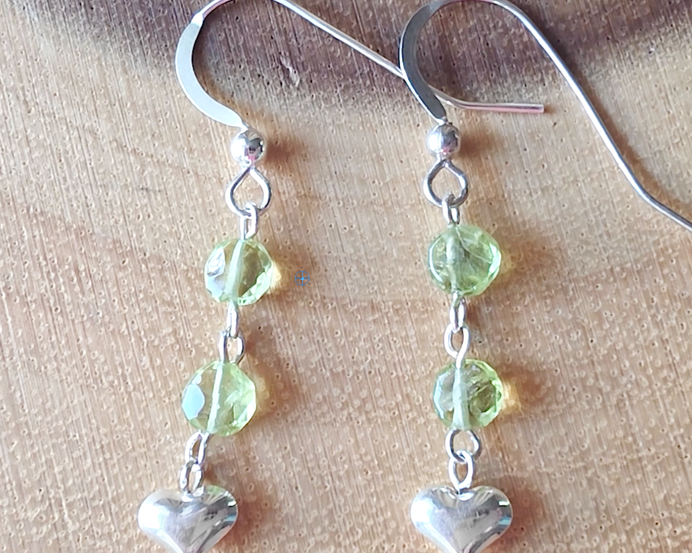 A pair of long sparkly Peridot Sterling silver earrings with two round faceted green gemstones and silver hearts on the base dangling from french earring hooks on wood 