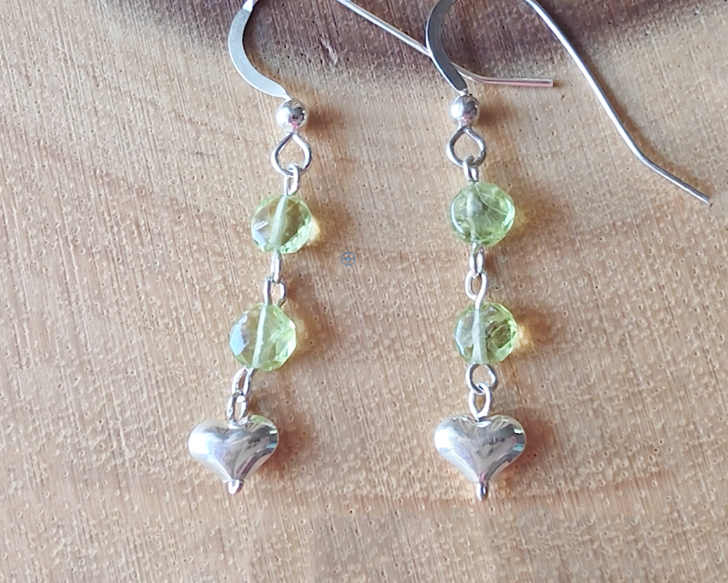 A pair of long sparkly Peridot Sterling silver earrings with two round faceted green gemstones and silver hearts on the base dangling from french earring hooks on wood 